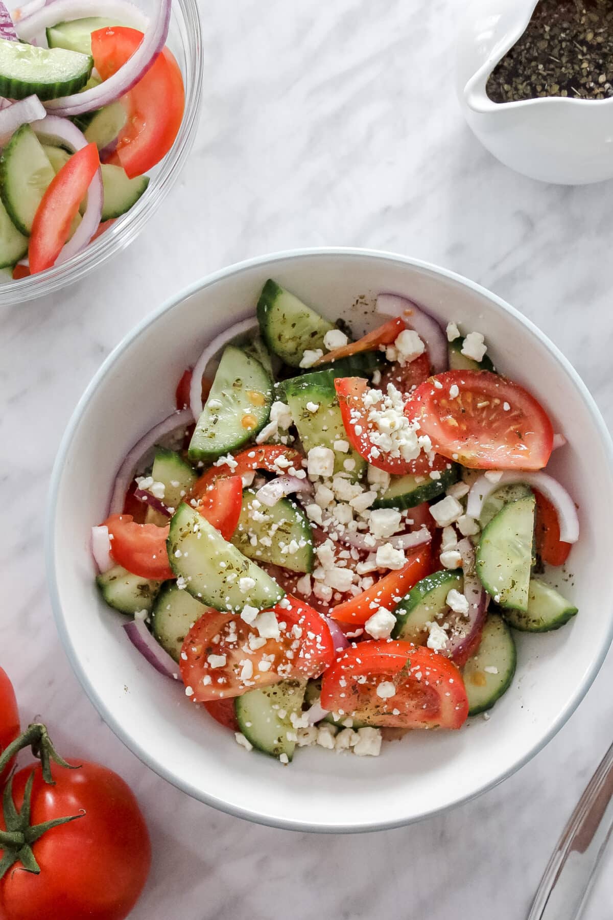 Overhead view of a bowl of Greek salad with tomatoes, cucumbers, red onions, and feta, next to a small pitcher of dressing.