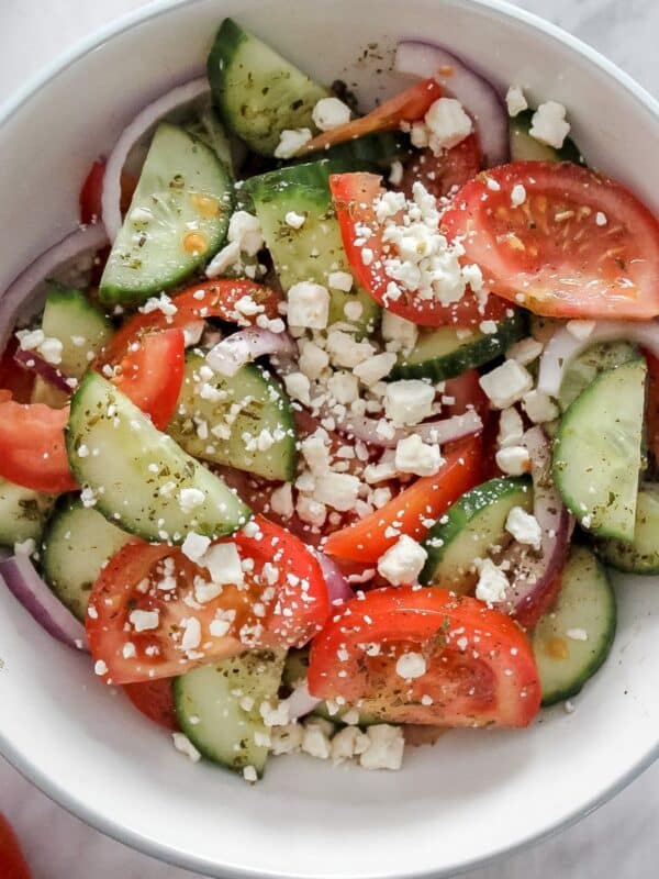 A bowl of Greek salad with cucumbers, tomatoes, red onions, olives, and crumbled feta, lightly tossed in vinaigrette.