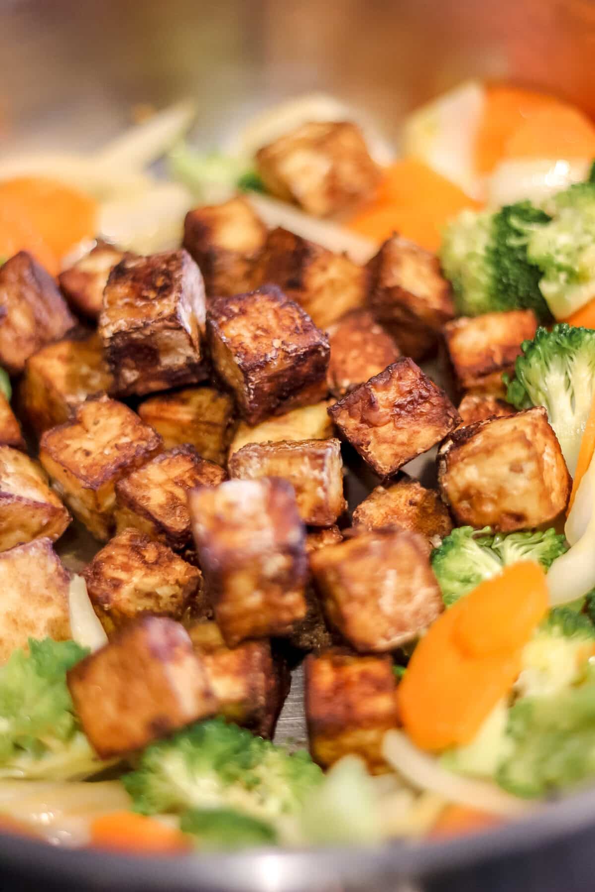 A close-up of crispy golden-brown tofu pieces in a pan with sautéed broccoli, carrots, and onions.