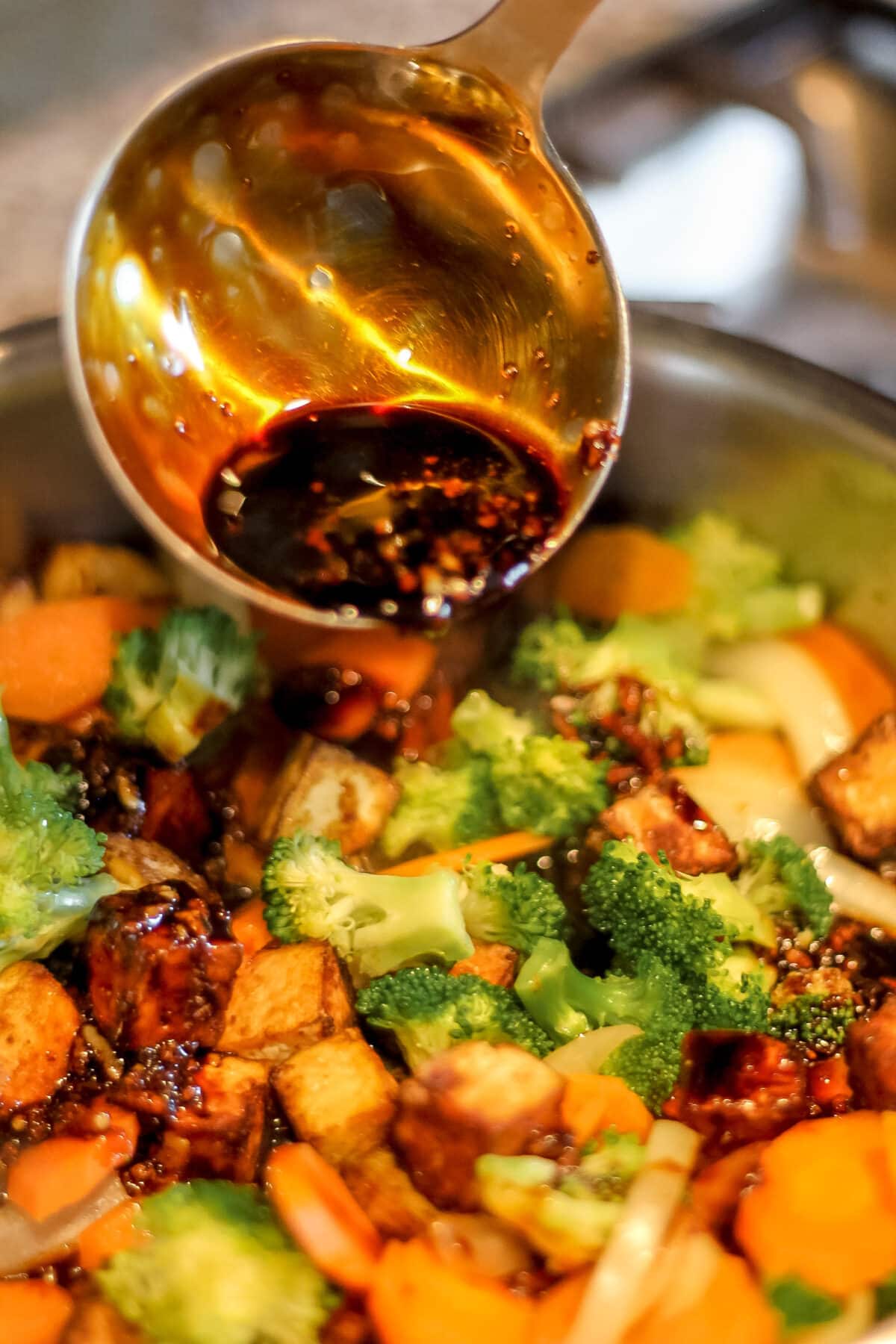 ladle pouring a rich, glossy stir-fry sauce over crispy tofu and vegetables in a pan.