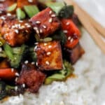 A close-up of crispy tofu stir fry coated in a glossy sauce, served over white rice with broccoli, red bell peppers, and sesame seeds, with chopsticks resting on the plate.