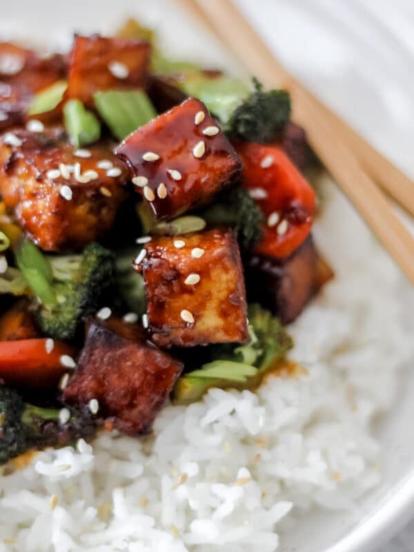 A close-up of crispy tofu stir fry coated in a glossy sauce, served over white rice with broccoli, red bell peppers, and sesame seeds, with chopsticks resting on the plate.