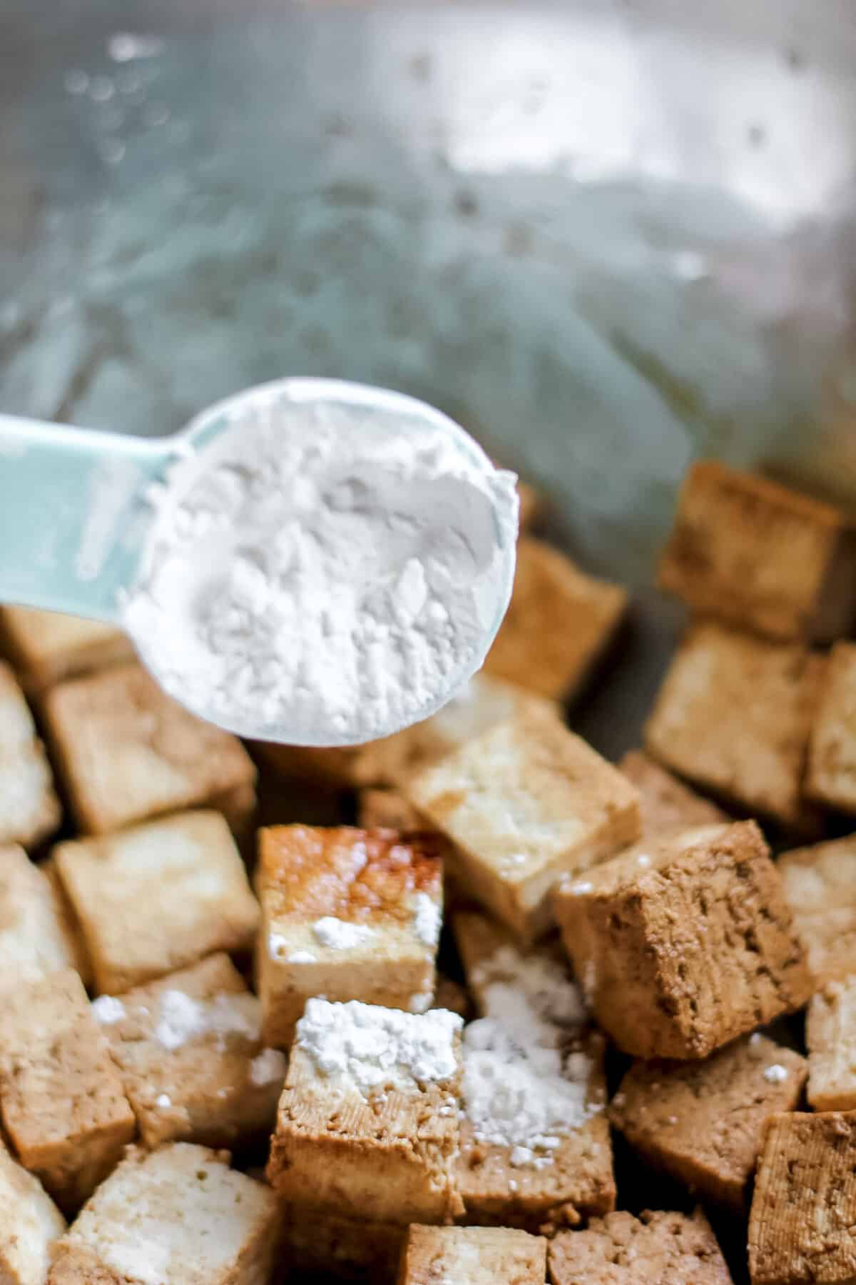 A spoonful of cornstarch being sprinkled over cubed tofu in a mixing bowl, preparing it for stir-frying.