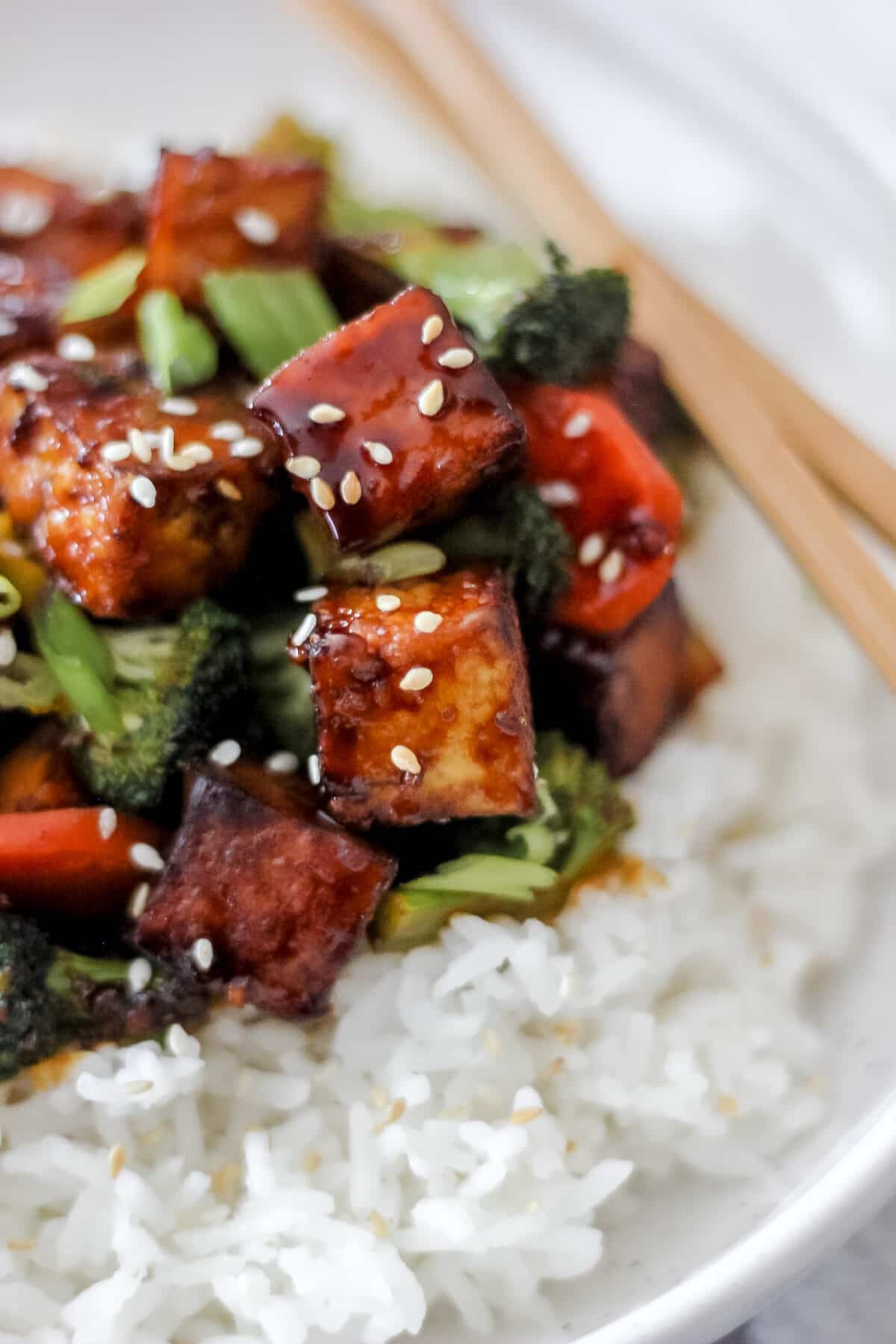 A close-up of crispy tofu stir fry coated in a glossy sauce, served over white rice with broccoli, red bell peppers, and sesame seeds, with chopsticks resting on the plate.