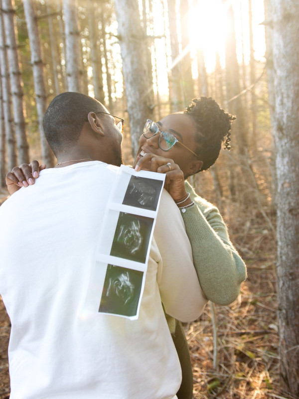 Husband and Wife Hugging in Woods with 3 Ultrasound Pictures of Baby