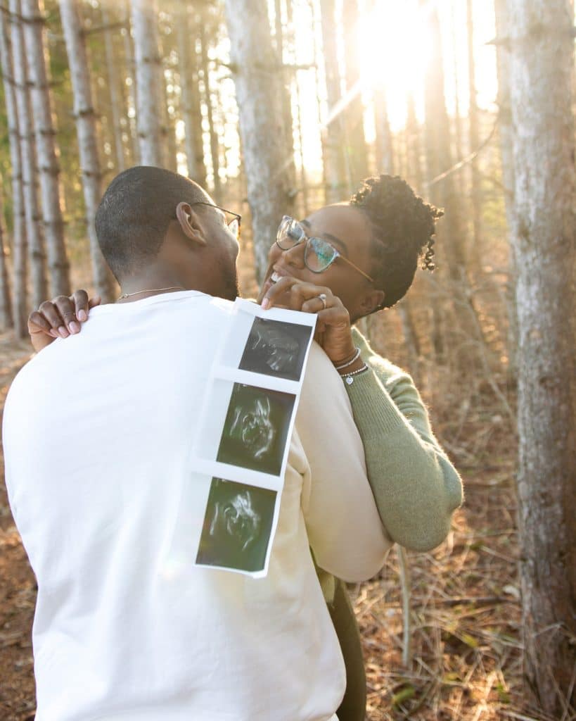 Husband and Wife Hugging in Woods with 3 Ultrasound Pictures of Baby
