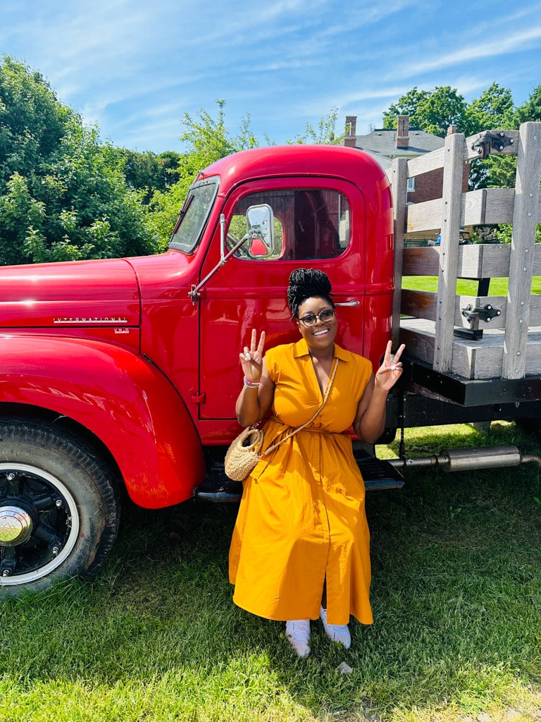 a woman in a yellow dress, smiling with both hands displaying peace signs sitting beside a bright red truck
