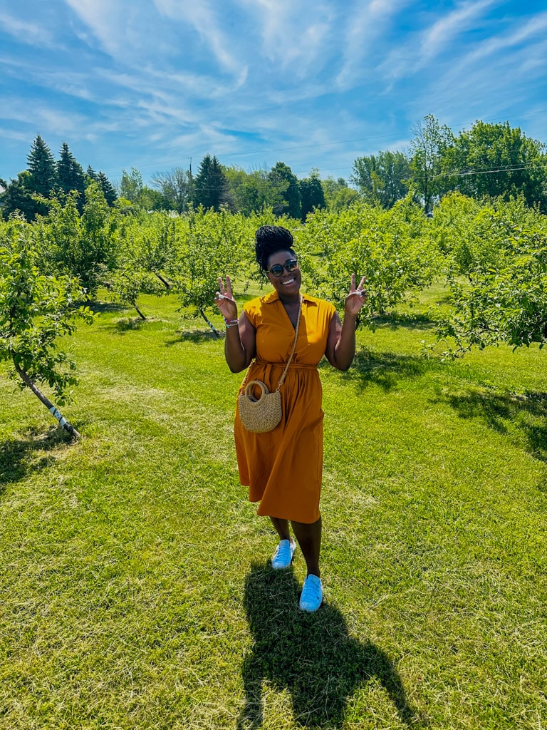 a woman in a yellow dress, smiling with both hands displaying peace standing in a vineyard in Prince Edward County