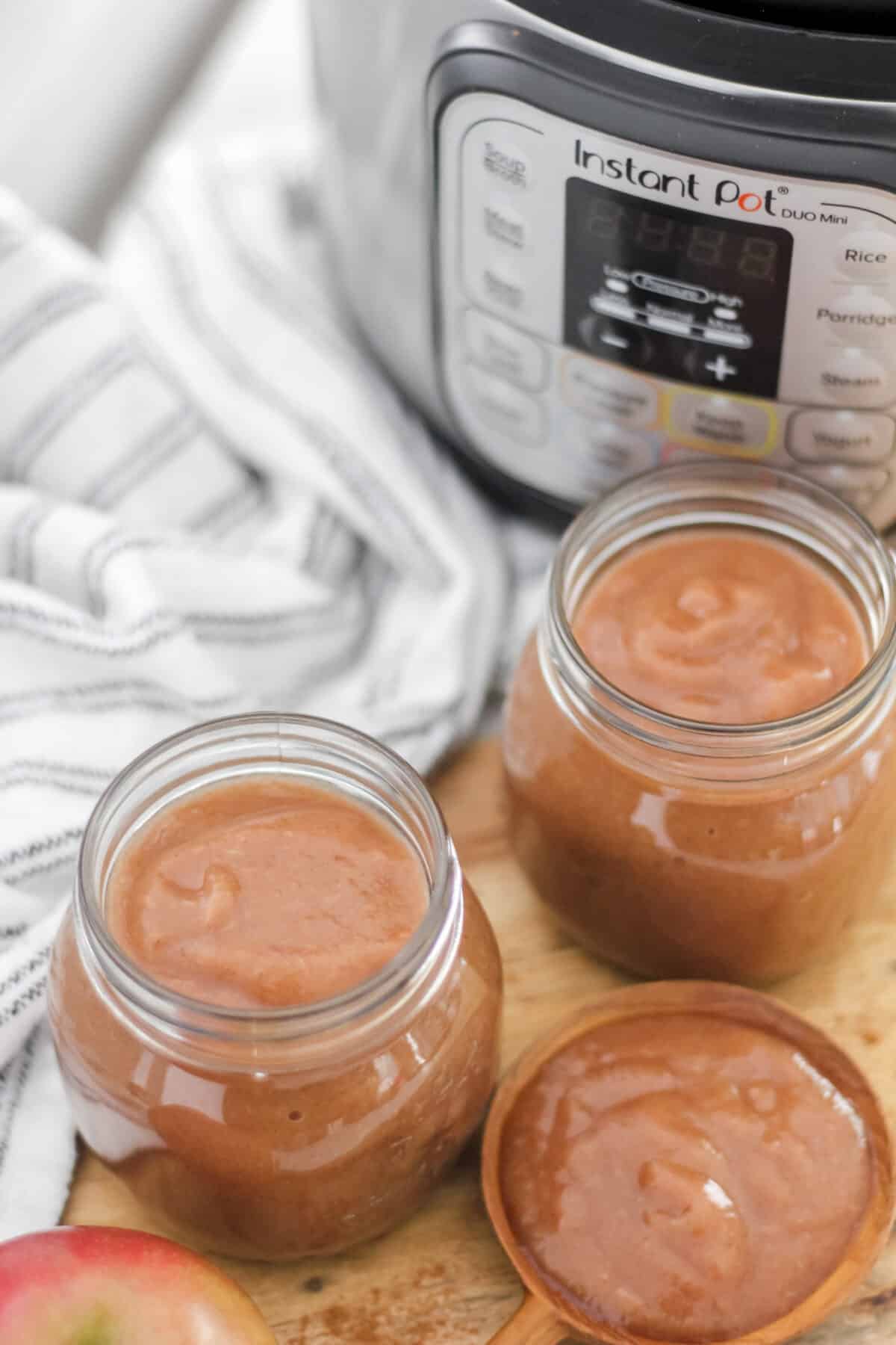 Two jars of apple butter next to a wooden spoon, with an Instant Pot and striped towel in the background.