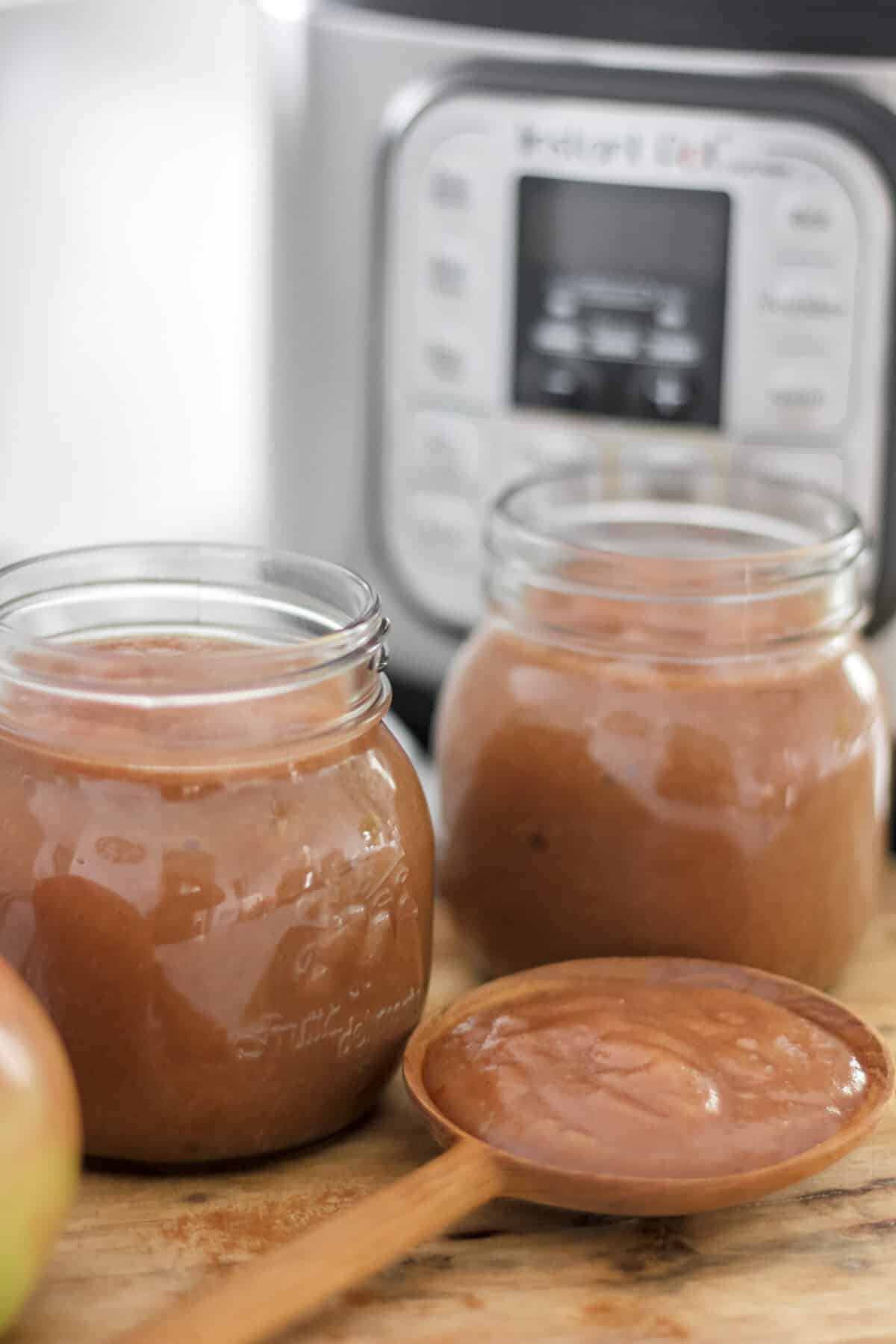 Jars of apple butter in front of an Instant Pot with a wooden spoonful of apple butter on a cutting board.