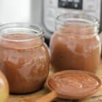 Two jars of smooth apple butter sit beside a wooden spoon filled with apple butter.