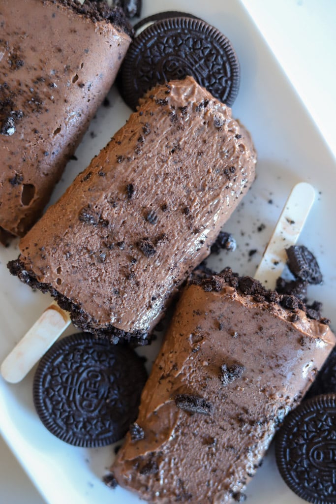 Chocolate cookies and cream pudding pops on a plate with crushed Oreo pieces and wooden sticks.
