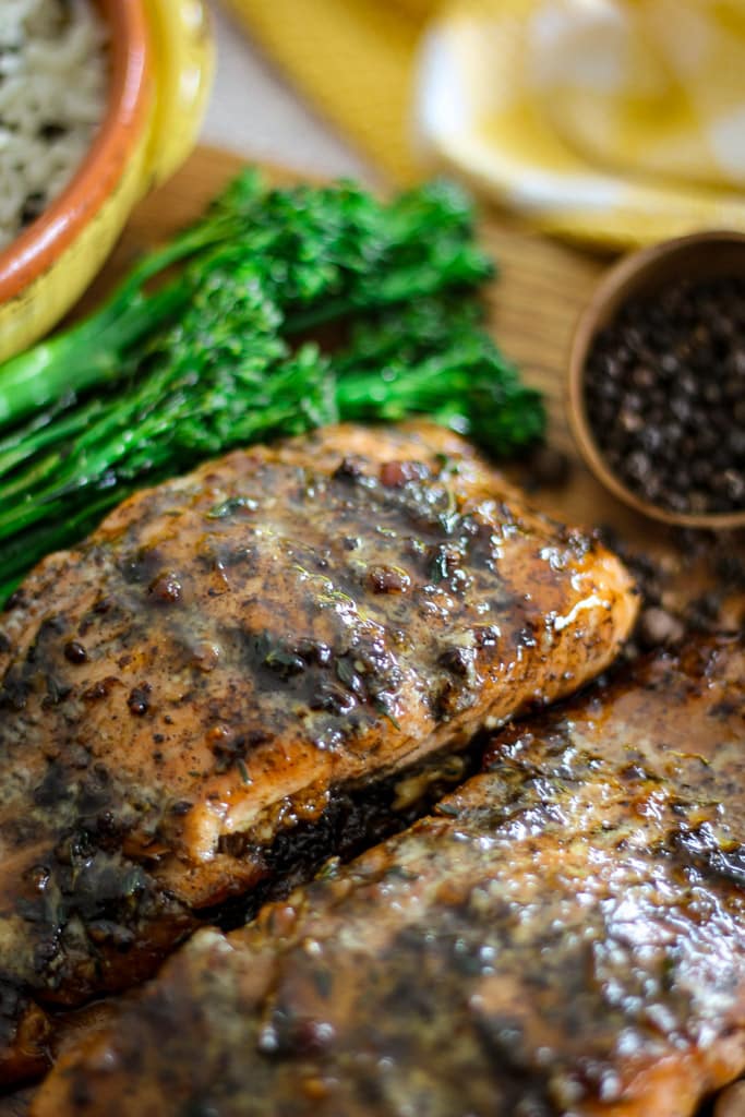 Close-up of glazed jerk salmon fillets with broccolini and a bowl of black peppercorns.