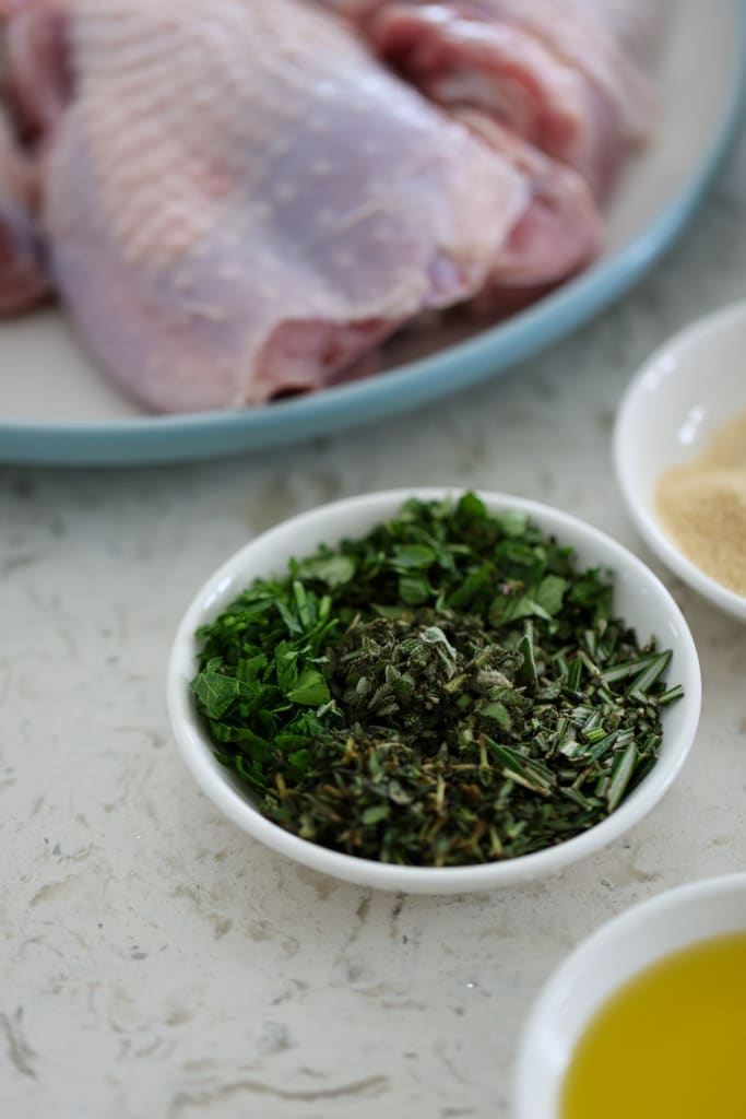 A small bowl of freshly chopped herbs beside raw turkey thighs.