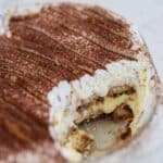A close-up of tiramisu in a glass dish with a large portion scooped out, showing layers of espresso-soaked ladyfingers, creamy filling, and cocoa topping.