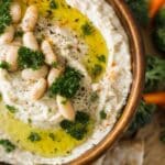 Bowl of creamy white bean dip topped with olive oil, parsley, and beans, served with pita chips.