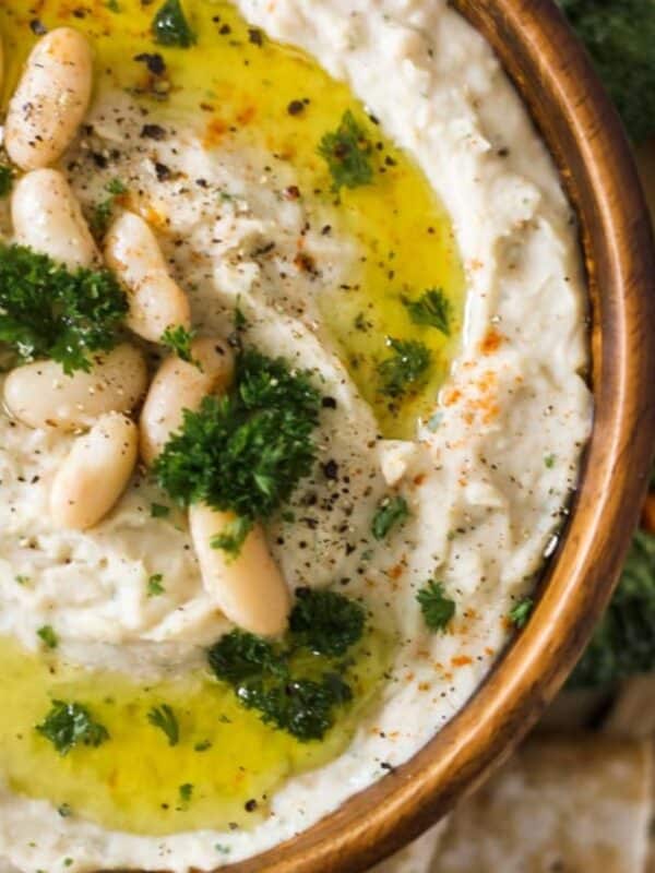 Bowl of creamy white bean dip topped with olive oil, parsley, and beans, served with pita chips.