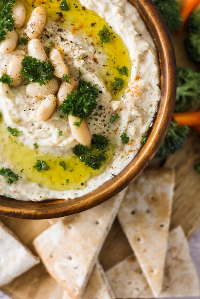 Bowl of creamy white bean dip topped with olive oil, parsley, and beans, served with pita chips.