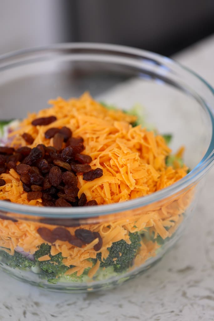 Close-up of shredded cheddar cheese and raisins on top of broccoli salad ingredients in a glass bowl.