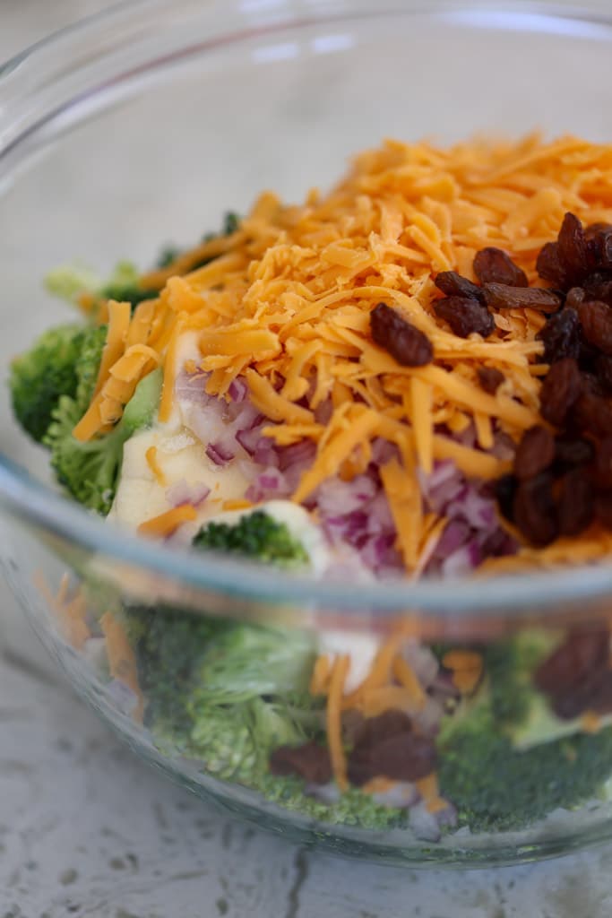Glass bowl with broccoli, cheddar cheese, red onion, and raisins before mixing.