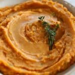 Overhead view of smooth sweet potato mash in a white bowl with thyme garnish.
