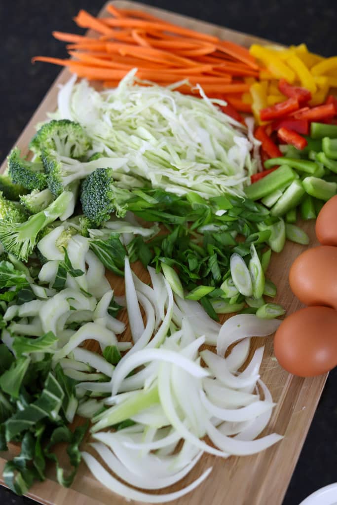 A cutting board with chopped vegetables including broccoli, cabbage, onions, and green beans beside whole eggs.
